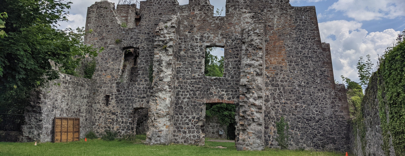 Ruinöses Mauerwerk aus Basaltbruchsteinen etwa drei Stockwerke hoch mit leeren Fensterlöchern. Links und rechts von Mauern eines Hofareals begrenzt. (2/3)
