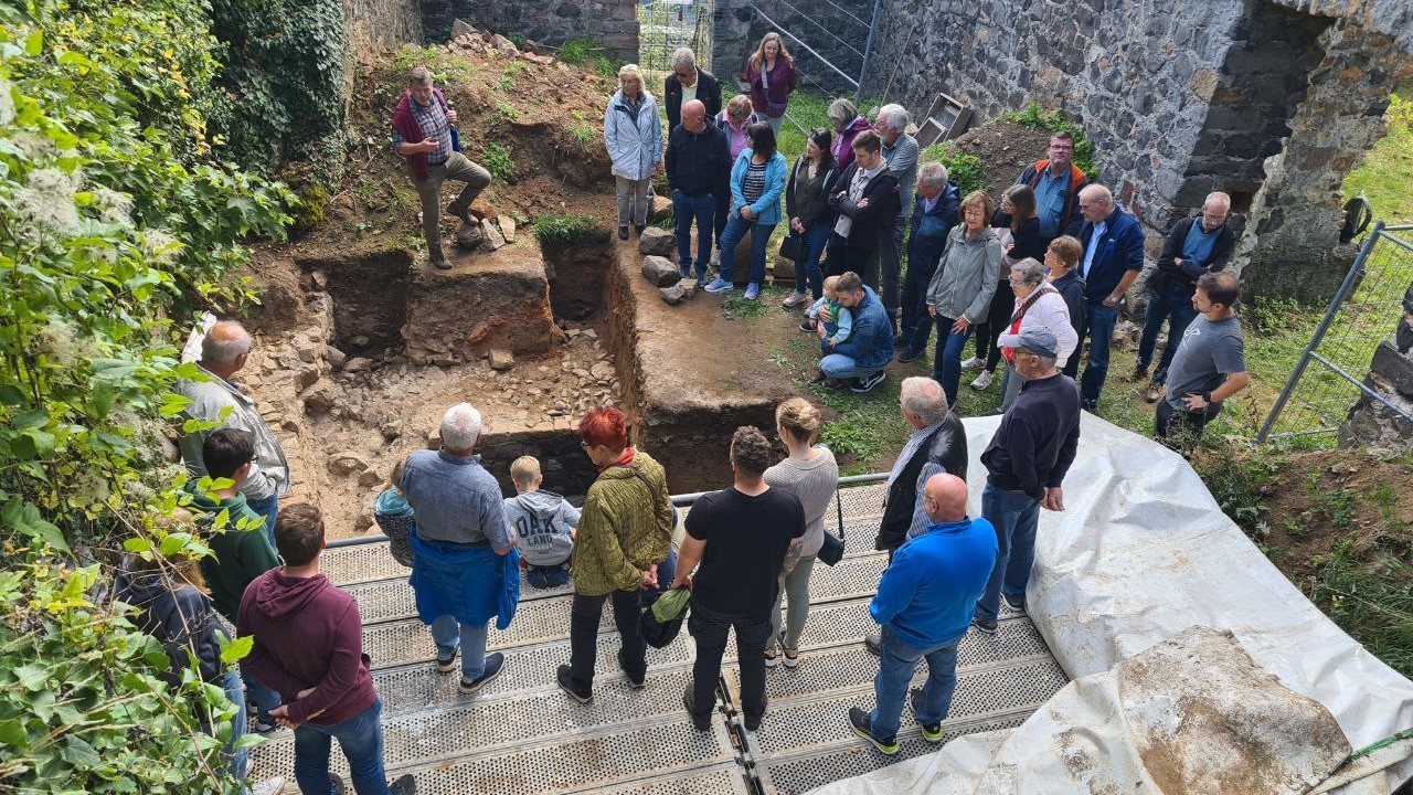 Eine große Gruppe Menschen umsteht eine archäologische Grabung in einer historischen Ruine. Eine Person erläutert Grabungsbefunde.